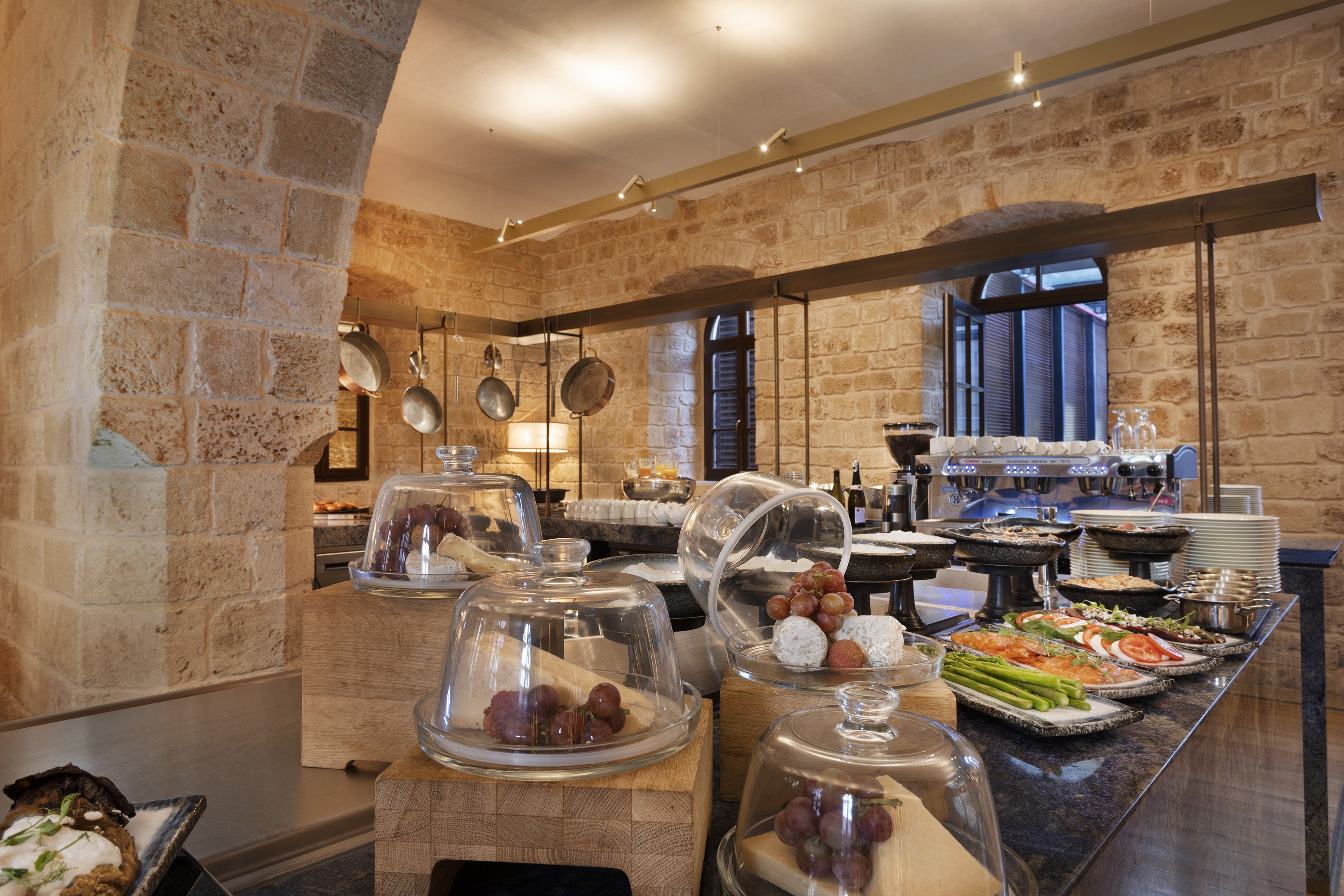 Un bufé con diversos alimentos, entre ellos frutas, quesos y verduras, en una sala con paredes de piedra y ventanas amplias.