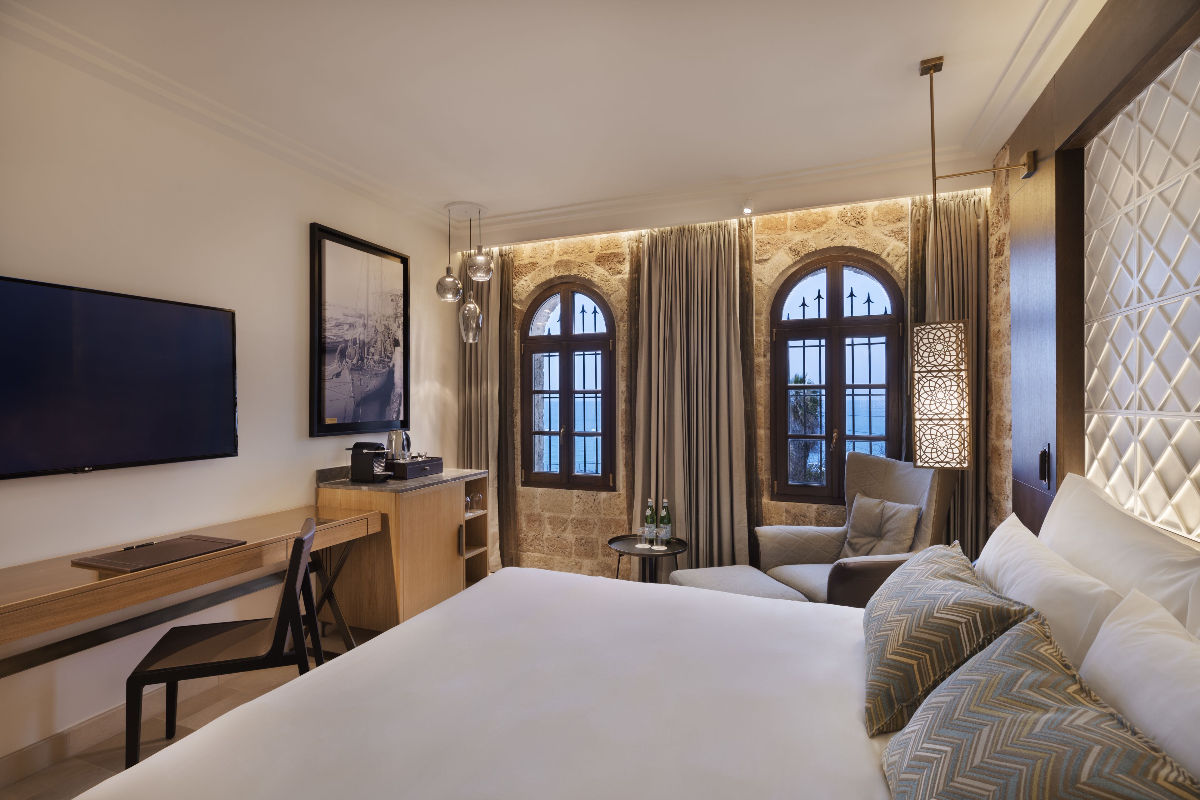 Habitación de hotel con una cama grande, un escritorio, una silla, un televisor y dos ventanas con cortinas.