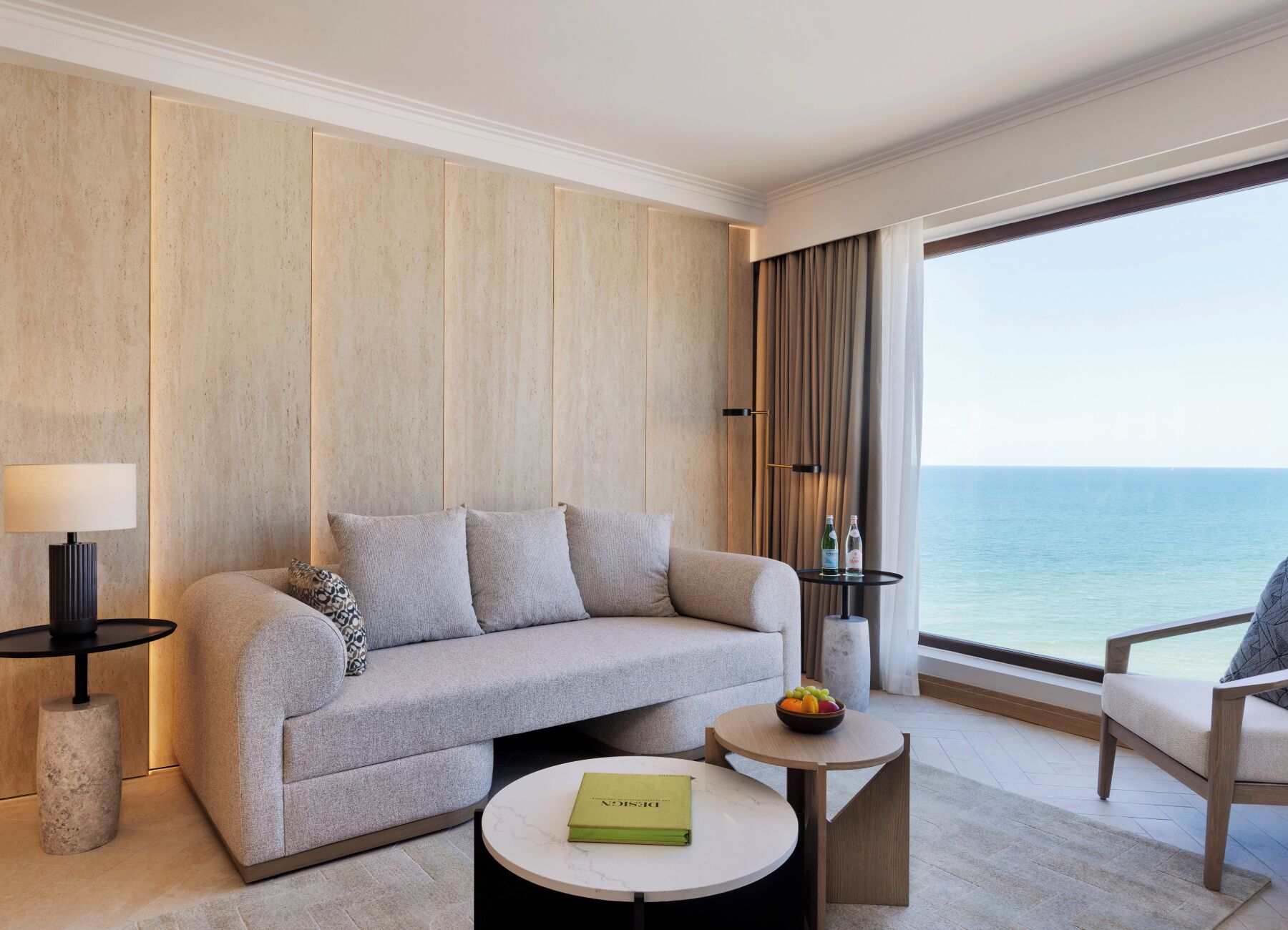 Habitación de hotel con sofá, una mesita redonda con un libro, una mesita auxiliar con lámpara y vista al mar.