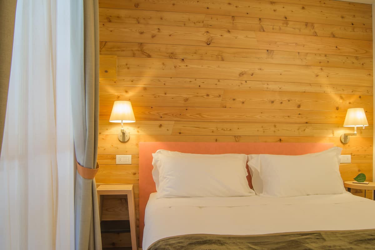 Une chambre d’hôtel avec une tête de lit en bois, deux lampes de chevet, un lit avec du linge de lit blanc et une table de chevet avec une lampe.