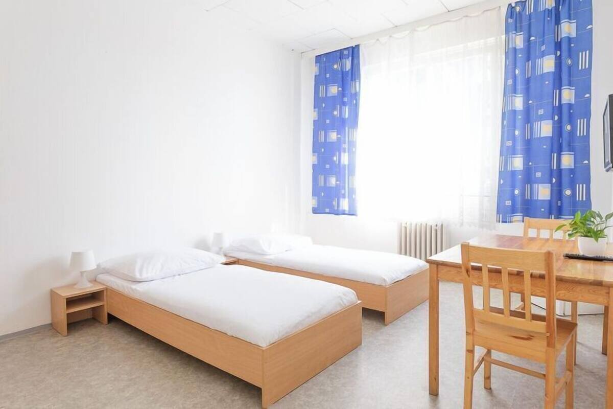 A room with two single beds, a wooden table with chairs, a small bedside table with a lamp, and a window with blue curtains.