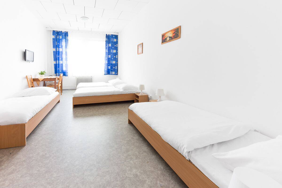 A modern hotel room with three single beds, a wooden desk, a chair, and a small table with a plant.