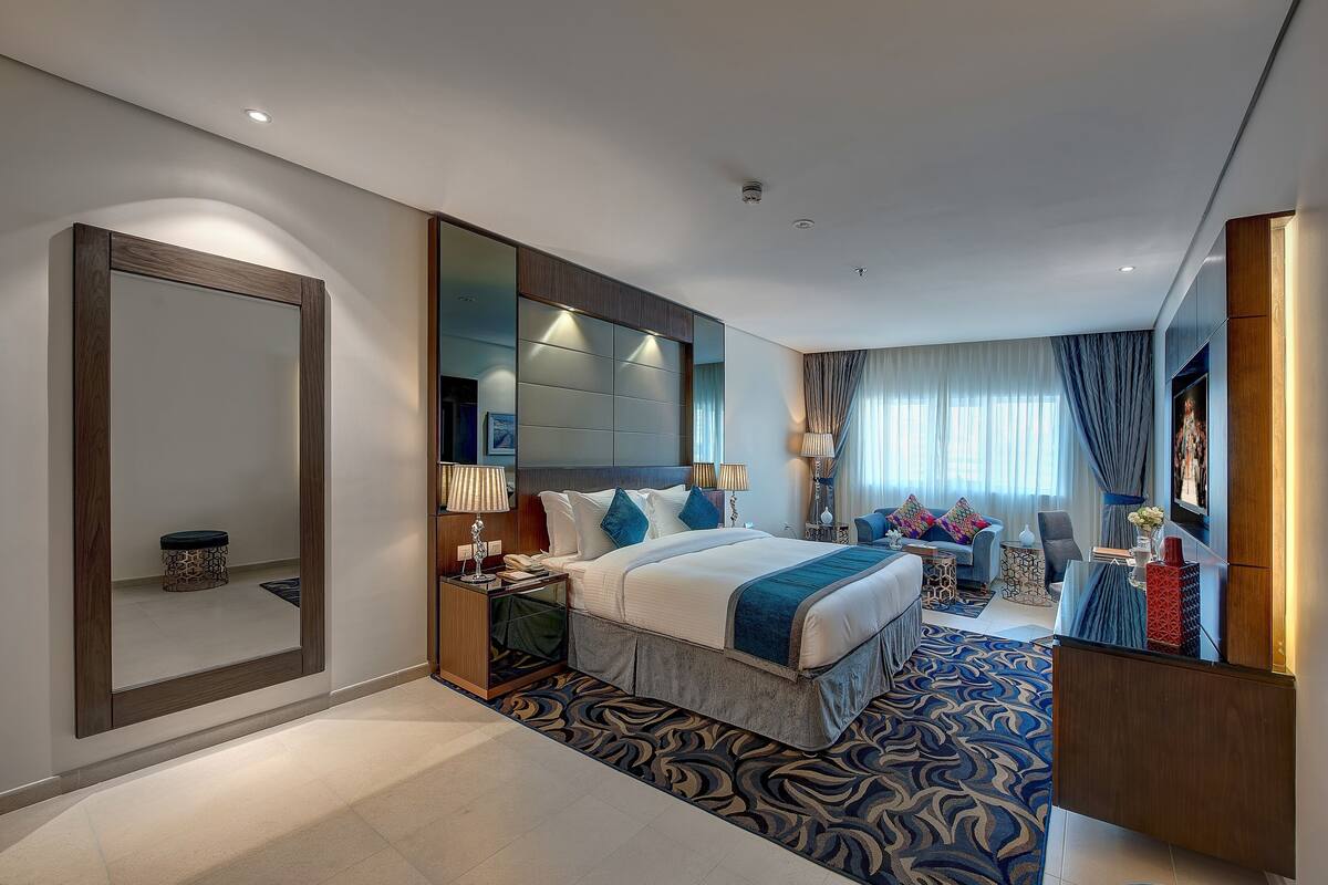 A modern hotel room with a large bed, a sitting area with a sofa and armchairs, a desk, and a large mirror.