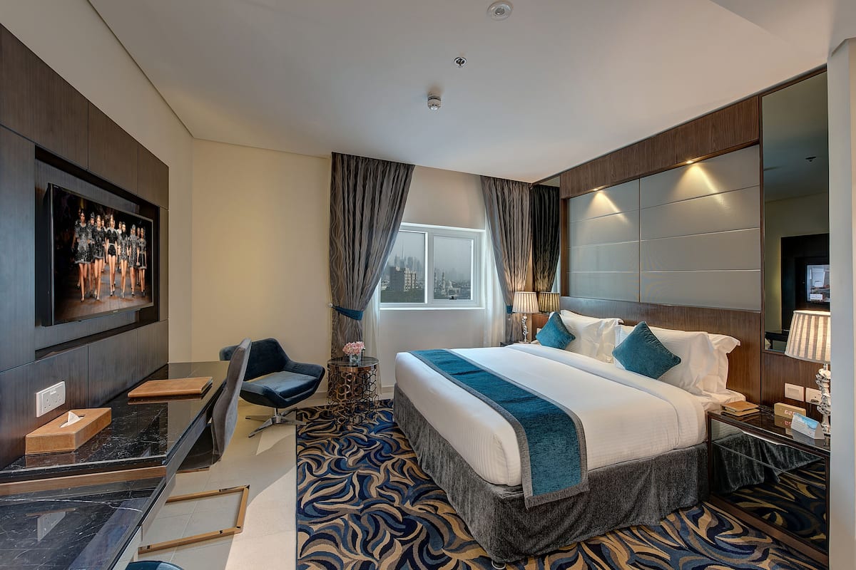 A modern hotel room with a large bed, a desk, a chair, a TV, and a view of the city.
