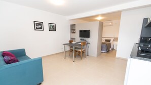 A modern living room with a blue sofa, a dining table with chairs, a kitchen area with a stove, and a bedroom with a bed and a TV. - Morada Clariana (Curitiba)