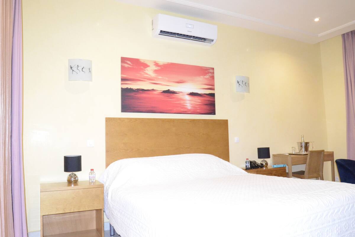 Ein Hotelzimmer mit einem Bett, einem Schreibtisch, einem Stuhl, einer Klimaanlage und einem Sonnenuntergangsbild.