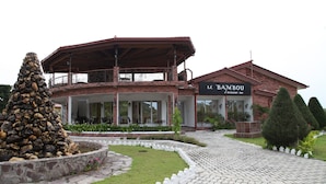 Un bar restaurante llamado "LE BAMBOU" con fachada de ladrillo, patio cubierto y jardín ajardinado con una fuente de rocas y arbustos cuidadosamente recortados.