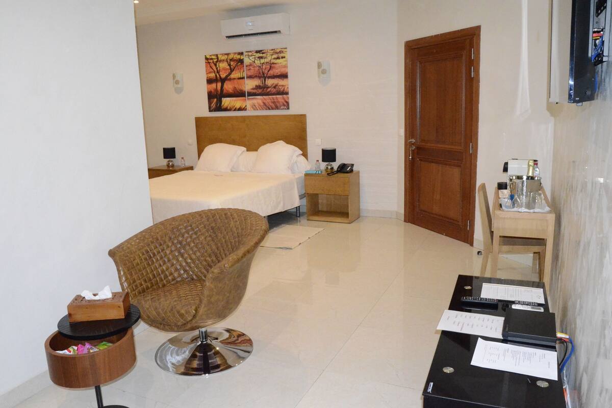 Ein Hotelzimmer mit einem Bett, einem Holzsessel, einem kleinen Tisch mit Papieren und einem an der Wand befestigten Fernseher.