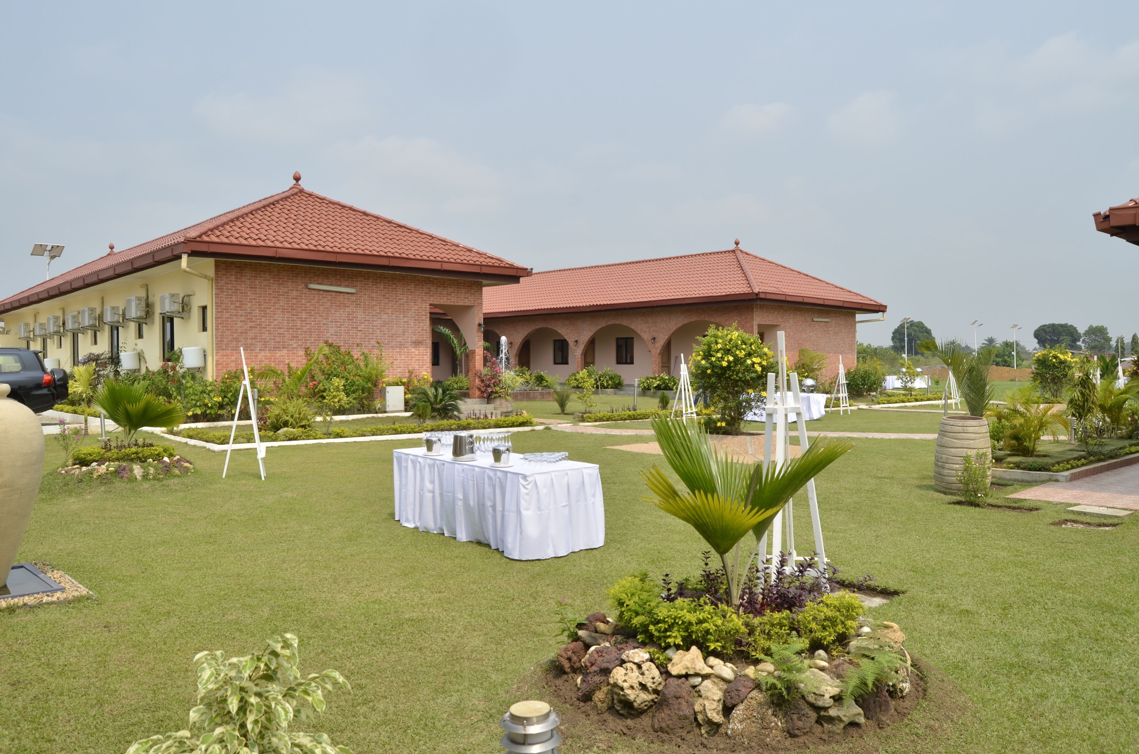 A building with a red-tiled roof and brick walls, a lawn with tables set up, and various plants and trees.