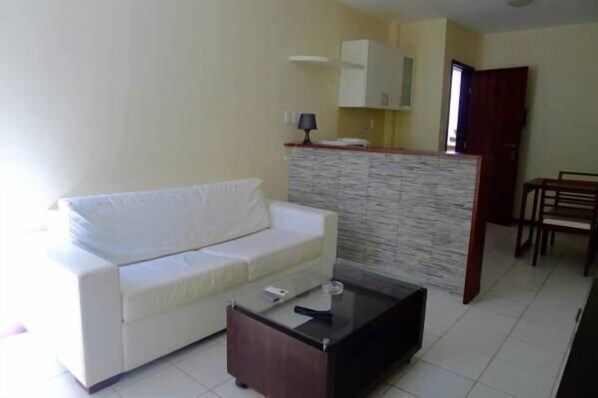 Un salón con sofá blanco, una mesa de centro de vidrio y una zona de cocina con salpicadero de piedra.