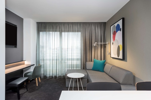 Una habitación de hotel moderna con un sofá gris, una pequeña mesa redonda, un escritorio con una silla y un ventanal con cortinas translúcidas.