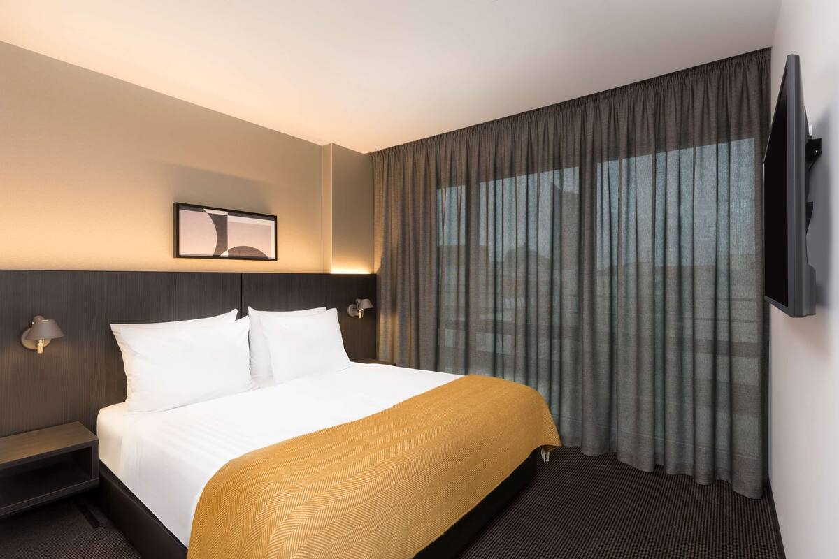 Ein Hotelzimmer mit Bett, Nachttisch, einem an der Wand befestigten Fernseher und einem Fenster mit Vorhängen.