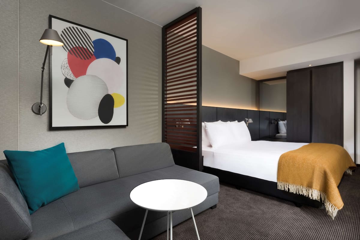 Ein modernes Hotelzimmer mit grauem Sofa, einem kleinen runden Tisch, einem Bett mit weißer Tagesdecke und einem senfgelben Überwurf, einem großen abstrakten Gemälde und einer Stehlampe.