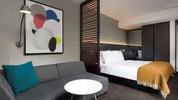 Een moderne hotelkamer met een grijze bank, een klein rond tafeltje, een bed met een witte deken en een mosterdkleurige plaid, een groot abstract schilderij, en een vloerlamp.