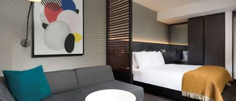 Ein modernes Hotelzimmer mit grauem Sofa, einem kleinen runden Tisch, einem Bett mit weißer Tagesdecke und einem senfgelben Überwurf, einem großen abstrakten Gemälde und einer Stehlampe.
