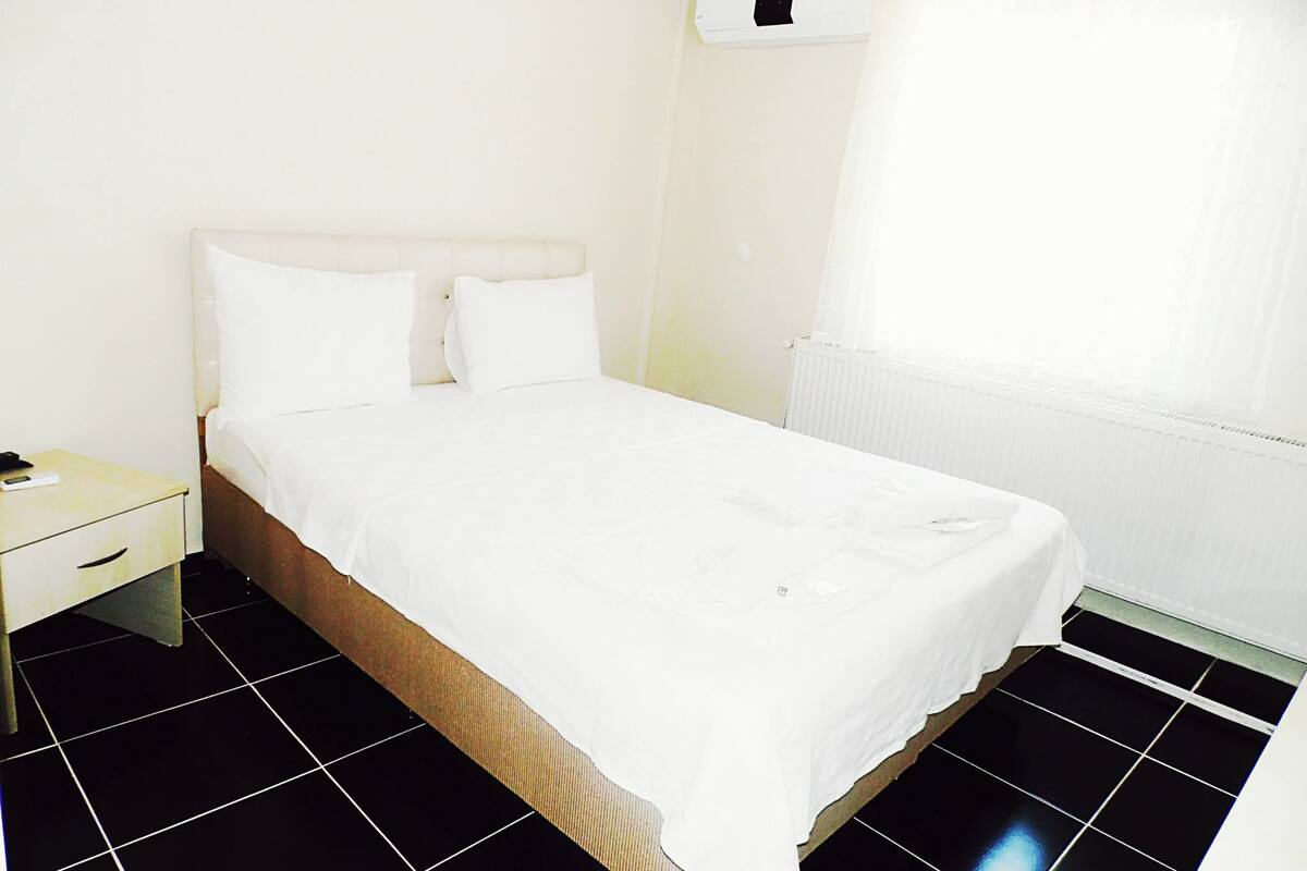 A hotel room with a bed, a nightstand, a white wall, a window with sheer curtains, and a radiator.
