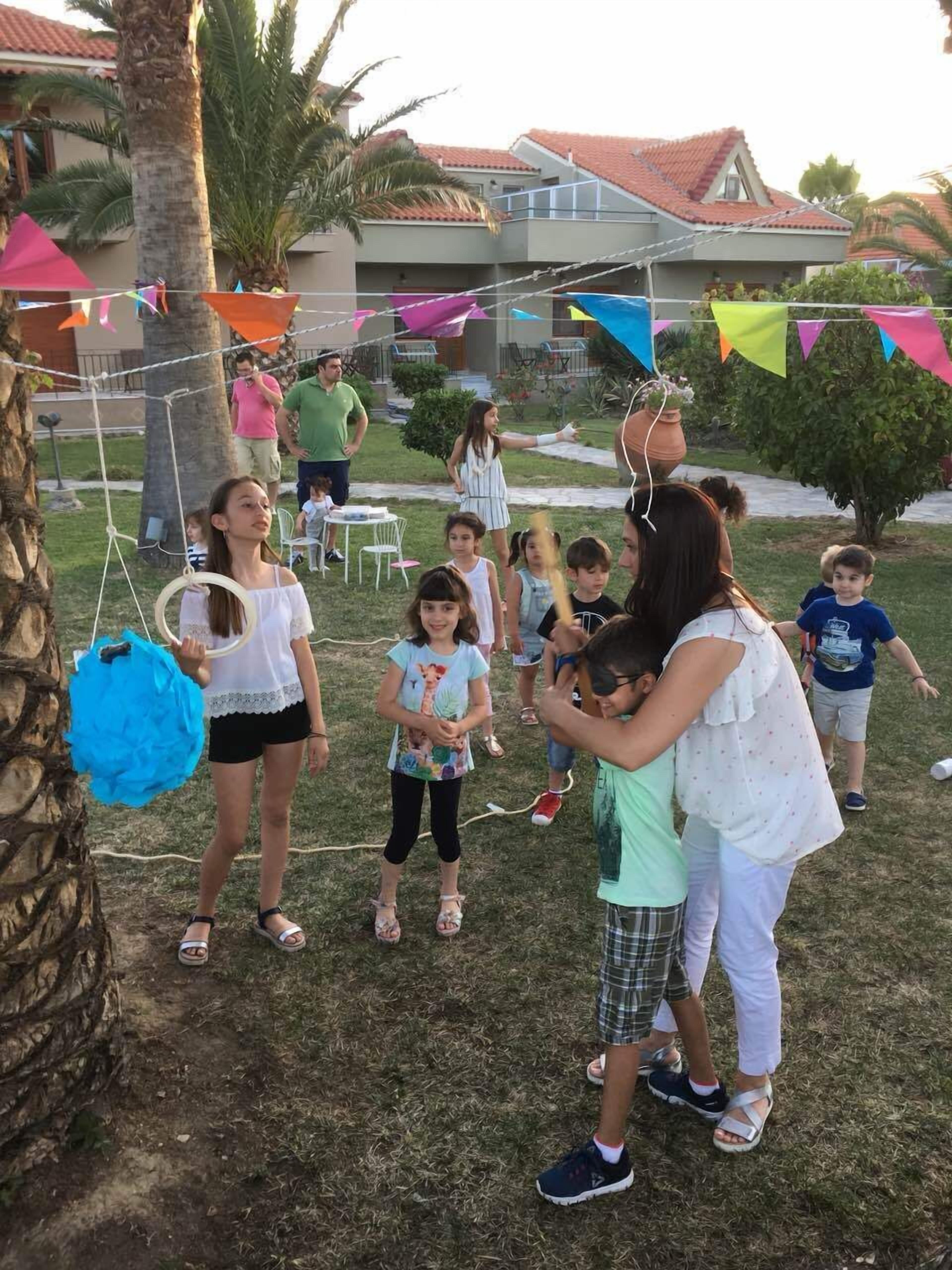 A group of children playing with a piñata in a grassy outdoor area with palm trees and a house in the background.