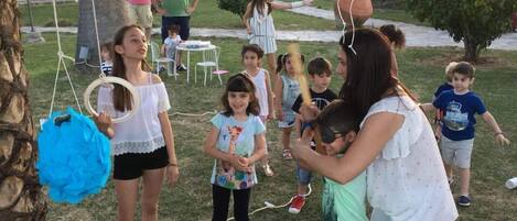 A group of children playing with a piñata in a grassy outdoor area with palm trees and a house in the background.