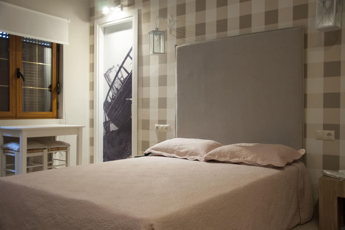 Un dormitorio con una cama grande, una mesita pequeña y una silla. Hay una ventana con persianas y una puerta que da a una escalera.
