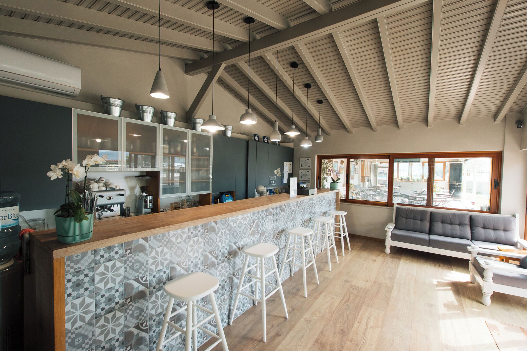 A modern kitchen with a bar area, white bar stools, a gray sofa, and large windows.