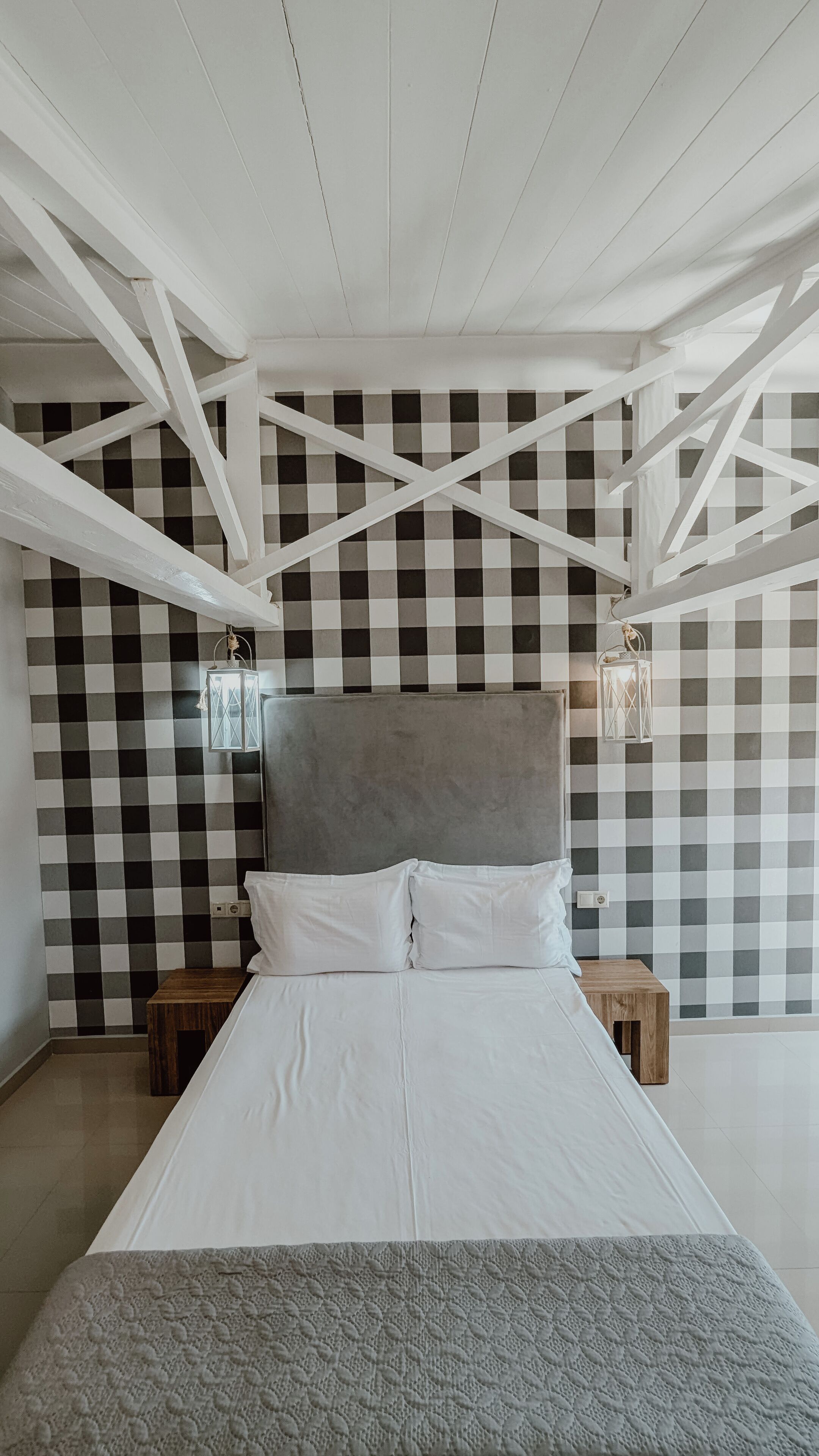 A bedroom with a checkered wall, a grey upholstered headboard, white bedding, and two bedside tables with lantern-style wall lights.