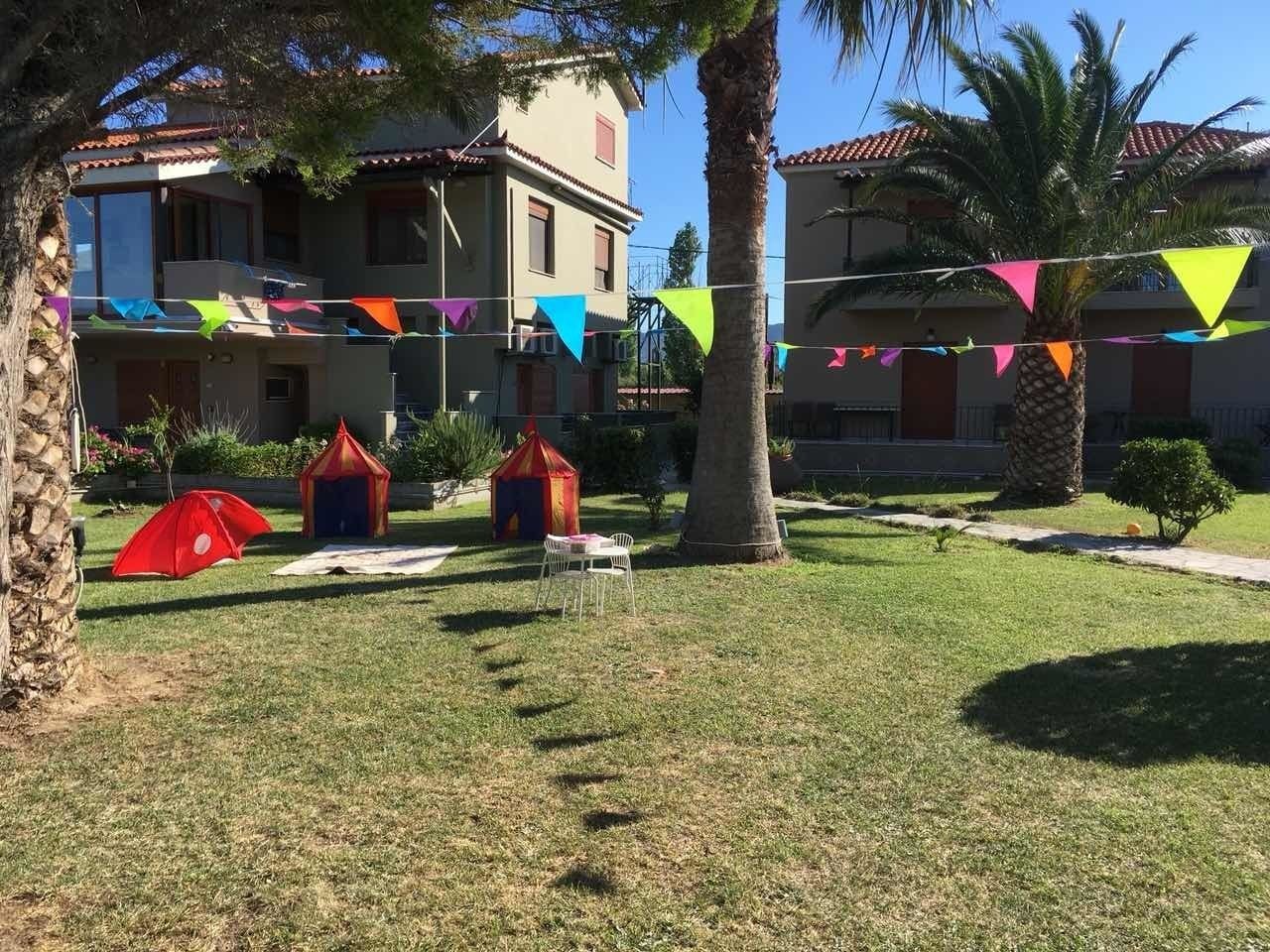 Une cour avec une pelouse verdoyante, des palmiers et des drapeaux triangulaires aux couleurs vives. On y trouve deux parasols rouges et une petite tente rouge et jaune.