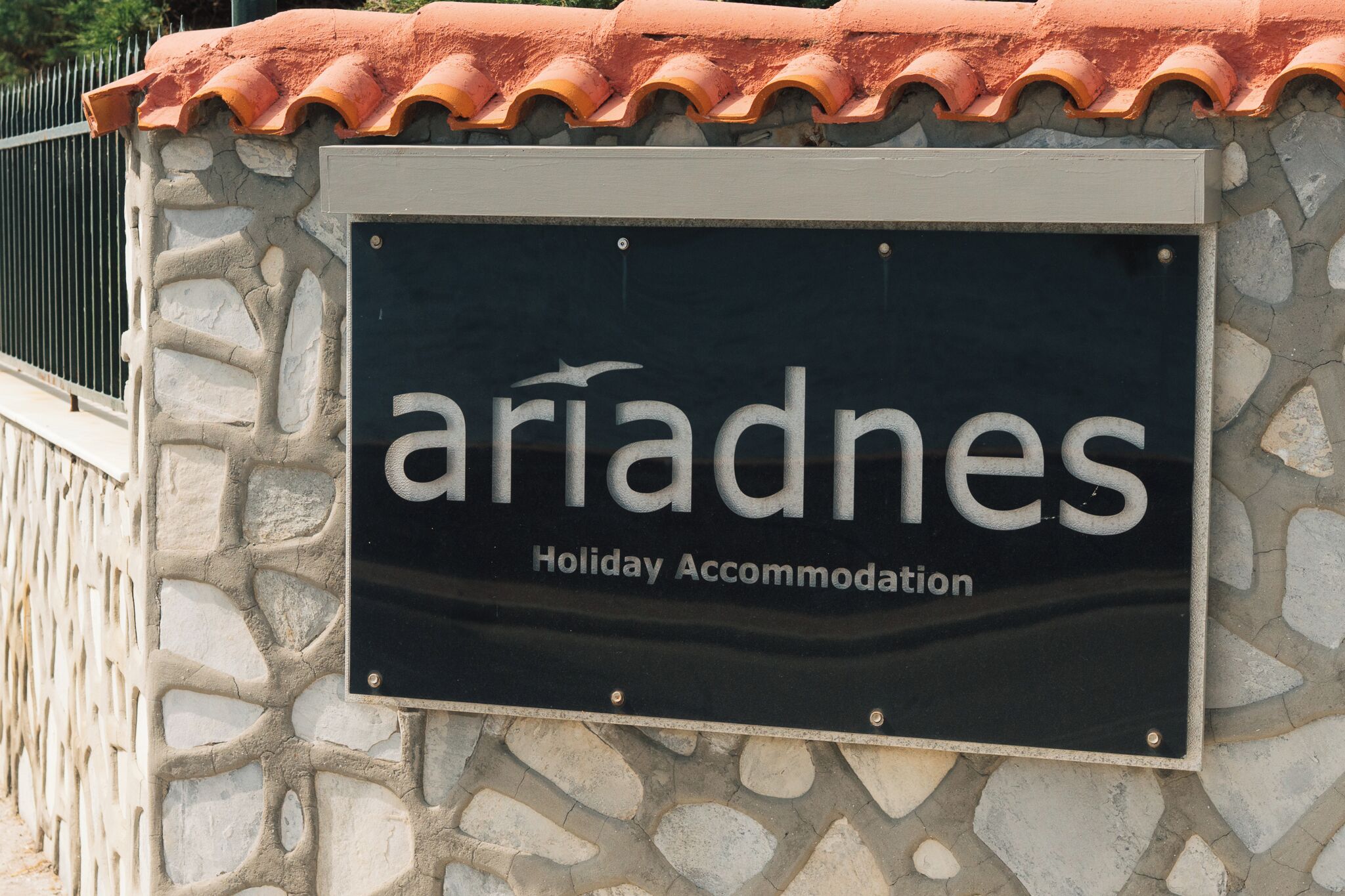 Une enseigne indiquant « Ariadnes Holiday Accommodation » est fixée à un mur de pierre surmonté d’un toit en tuiles.
