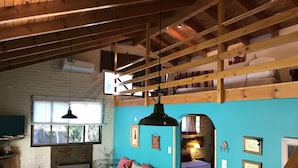 A cozy room with a wooden ceiling, a sofa with patterned cushions, a window with a view, and a wall with framed pictures. - El Colibri Cabañas de la Naturaleza (Trelew)