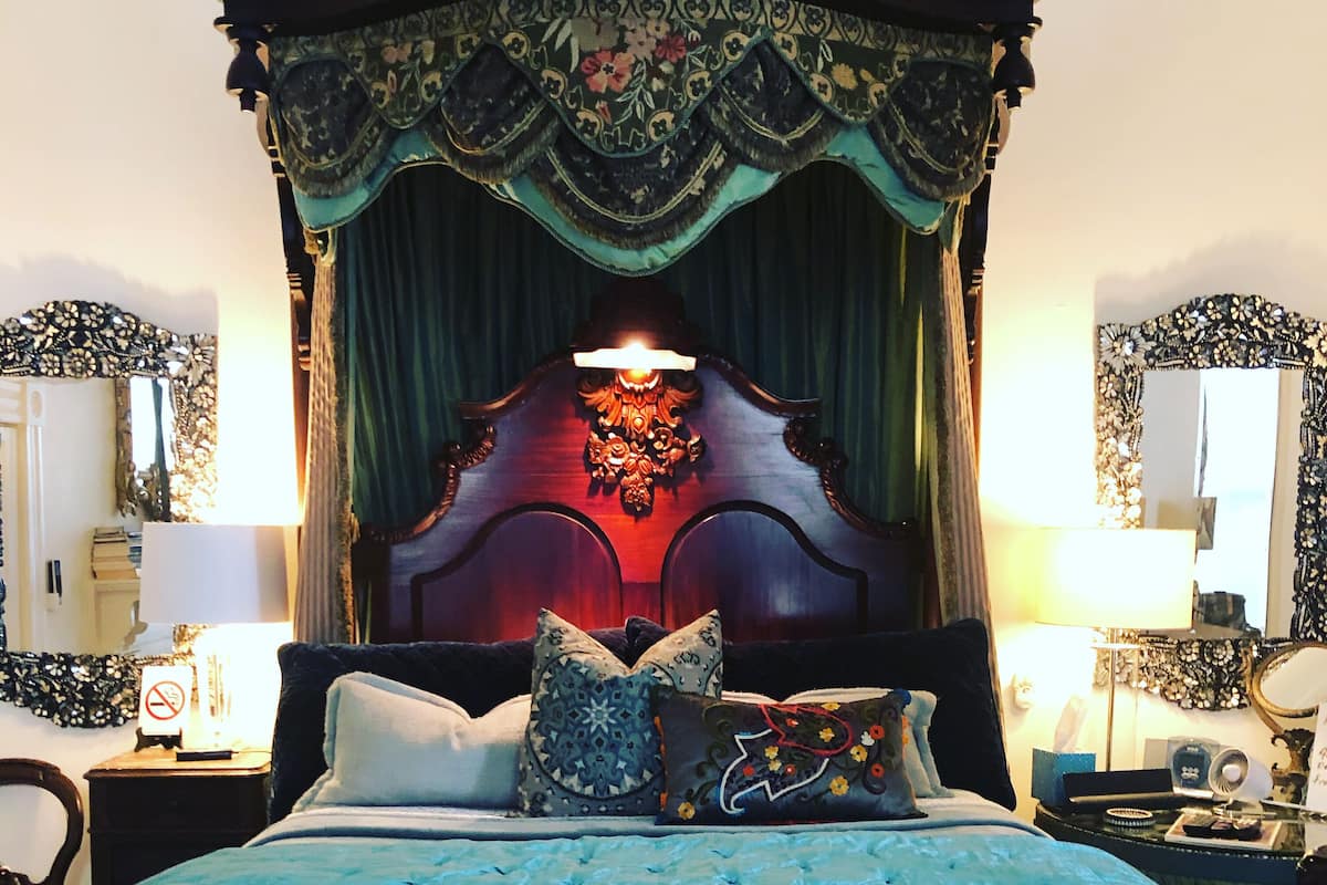 A bedroom with a four-poster bed, a bedside table with a lamp, and a mirror on the wall.