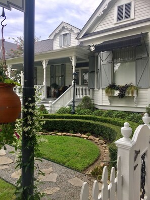 Una casa blanca con un porche, un jardín con seto y cerca de piquetes blancos.
