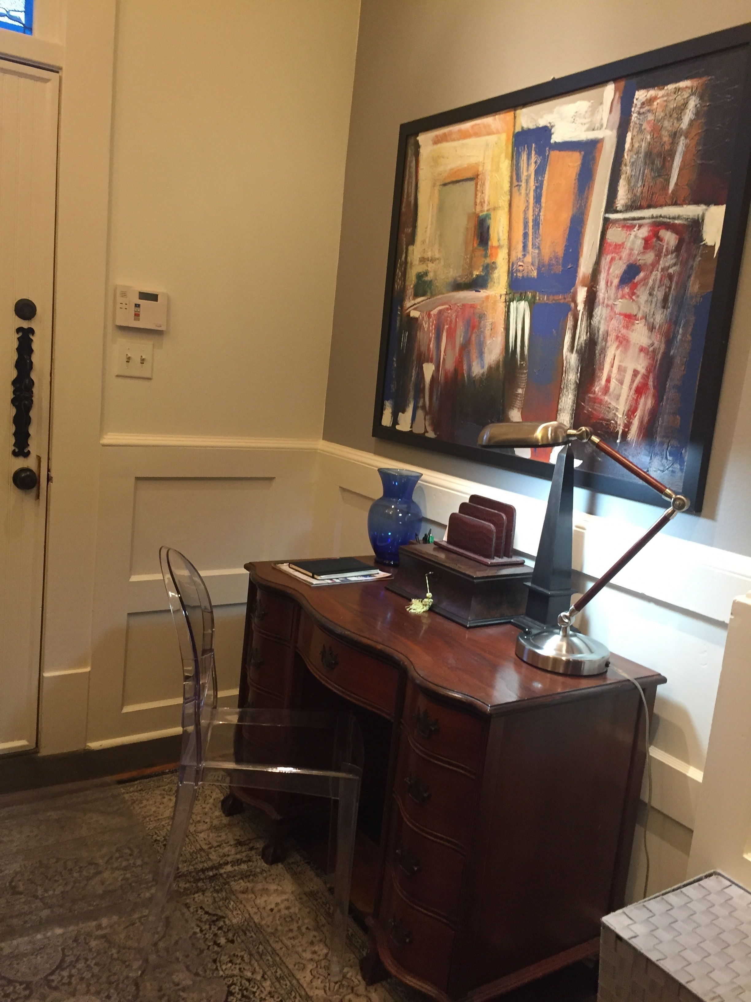 Un bureau en bois, une lampe, un vase bleu et un tableau abstrait encadré accroché au mur.