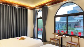 A hotel room with two beds, a small table with a basket and a glass, and a view of a building and trees through large windows. - Tuan Ngoc Hotel (Nam Hoa Lu)