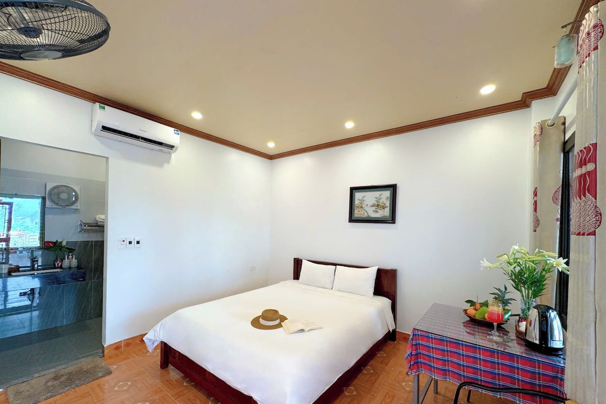 A bedroom with a bed, a small table with a vase, and a fan.