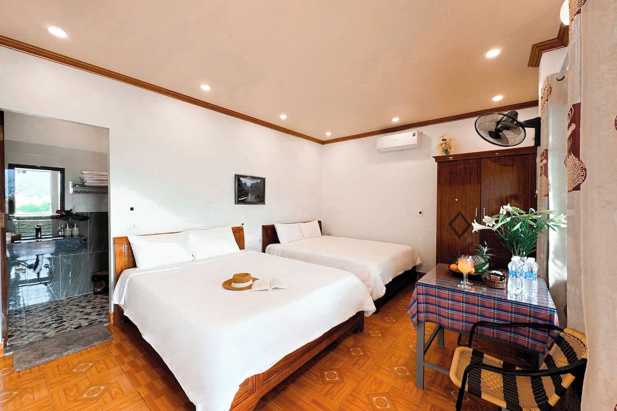 A hotel room with two beds, a dining table with a placemat, a fan, and a view of the kitchen.