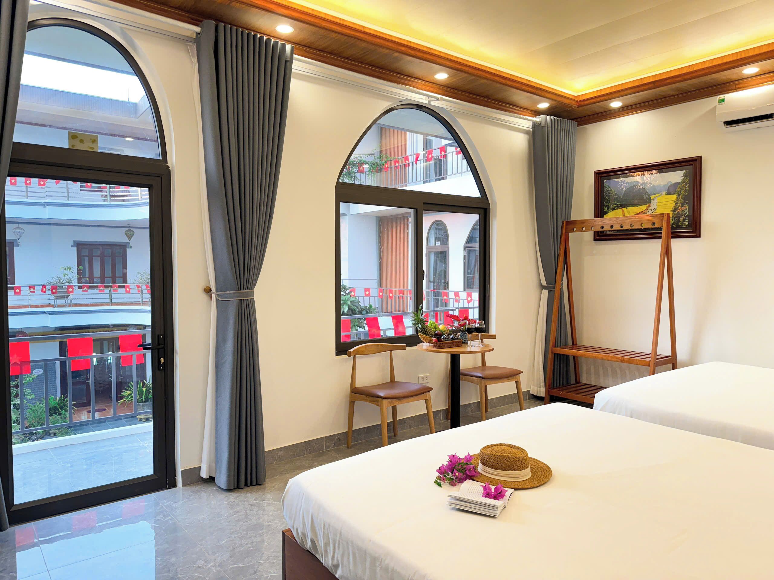 A hotel room with two beds, a dining table with chairs, a small balcony with red decorations, and a painting on the wall.