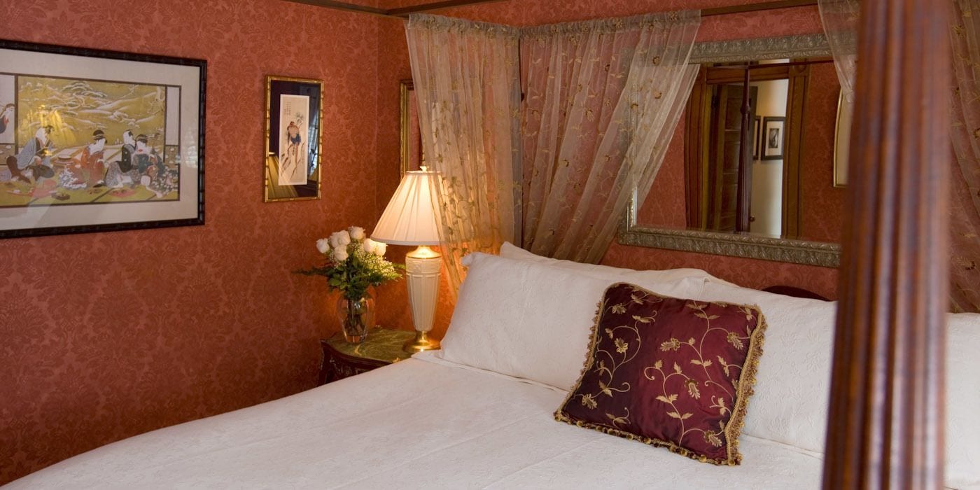 Habitación de hotel con cama, mesita de noche con lámpara, jarrón con flores e imagen enmarcada en la pared.