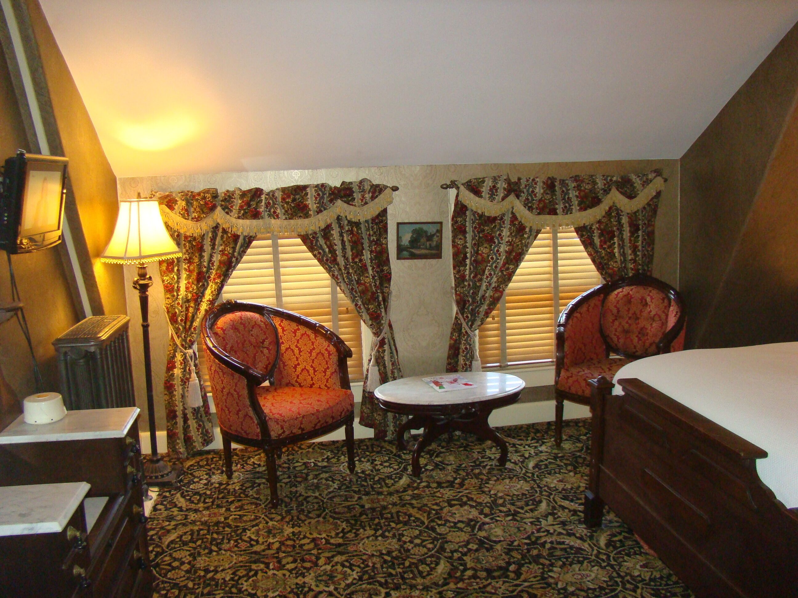 Een hotelkamer met een bed, twee fauteuils, een klein rond tafeltje, een lamp en een radiator.