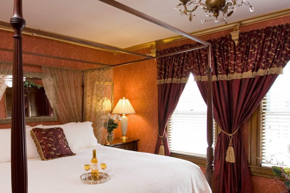 Una cama con dosel y mosquitera, una mesita de noche con lámpara, y una ventana con cortinas.