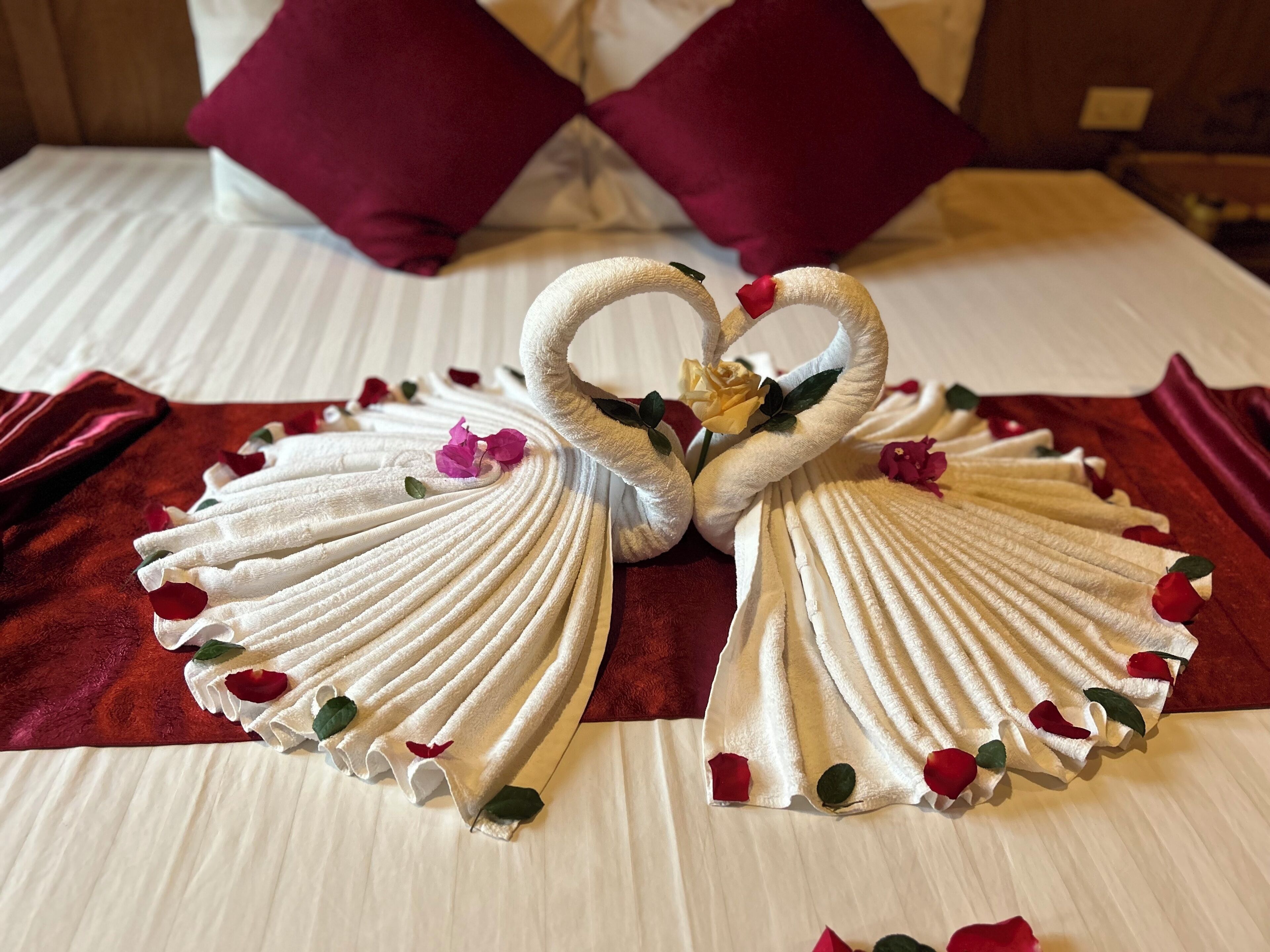 A bed with white linens, two red pillows, and a decorative swan made of towels with rose petals.