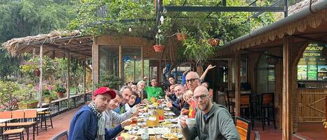 Um grupo de pessoas jantando em um restaurante ao ar livre, com vista para vegetação exuberante e montanhas ao fundo.
