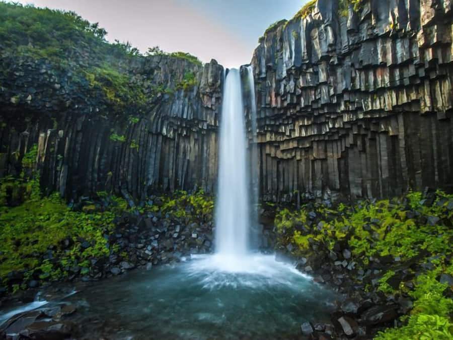 Uma cascata desce por um penhasco rochoso, rodeada por vegetação exuberante.