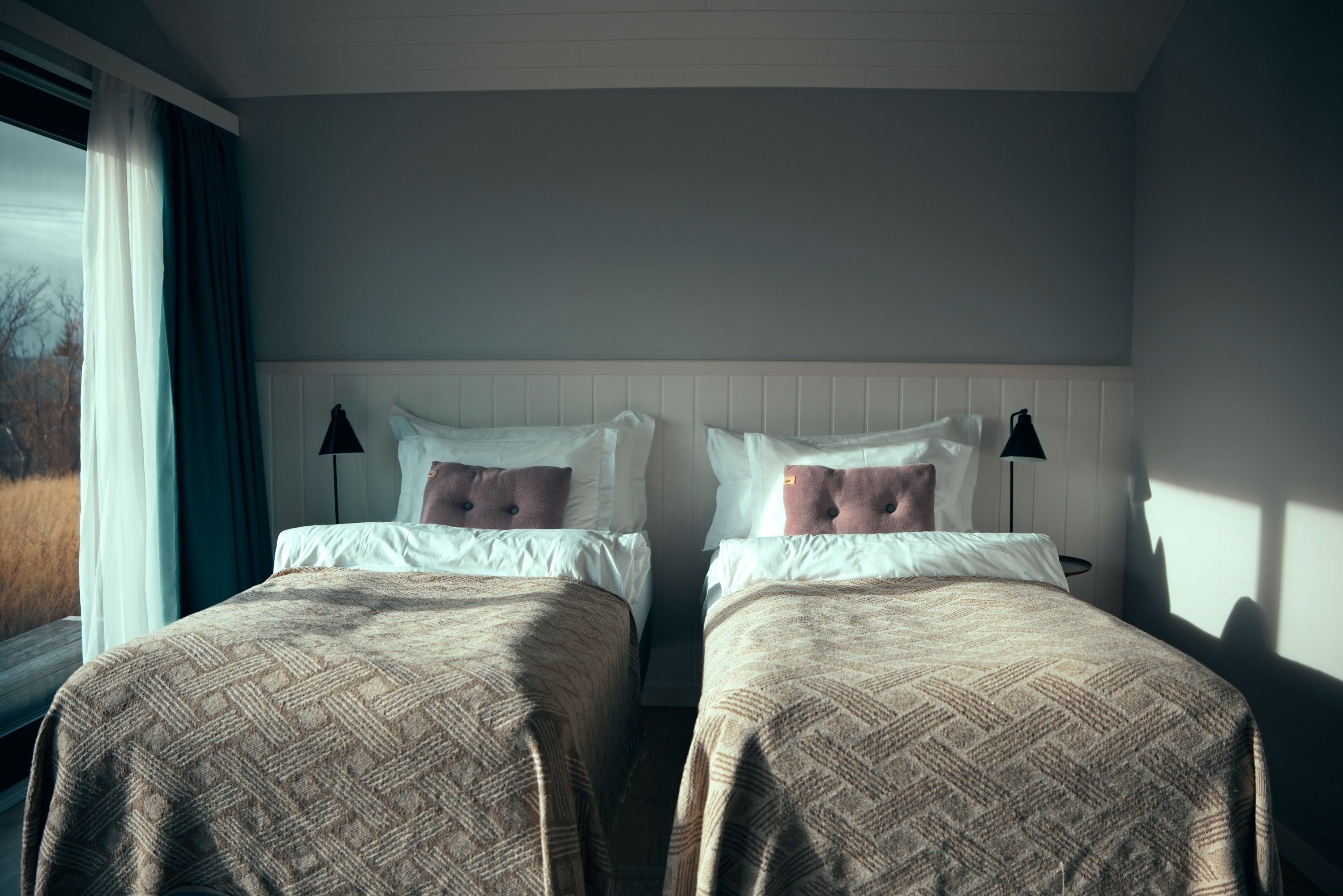 A room with two beds, each with a headboard, white bedding, and a patterned blanket. There are two bedside lamps on each bed.