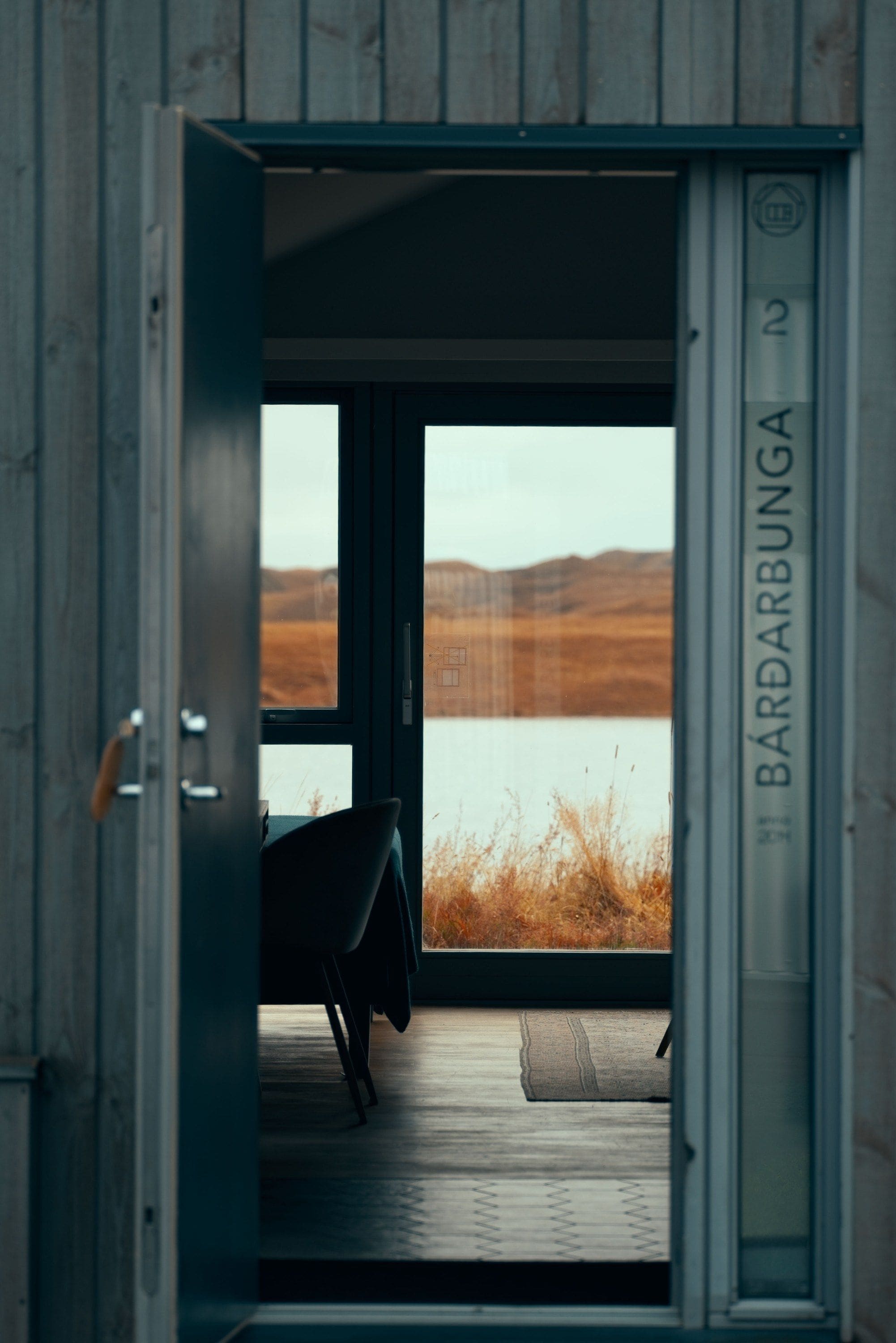 An open door leading to a room with a view of a lake and hills, a chair, and a door labeled 'BARBUNGA'.