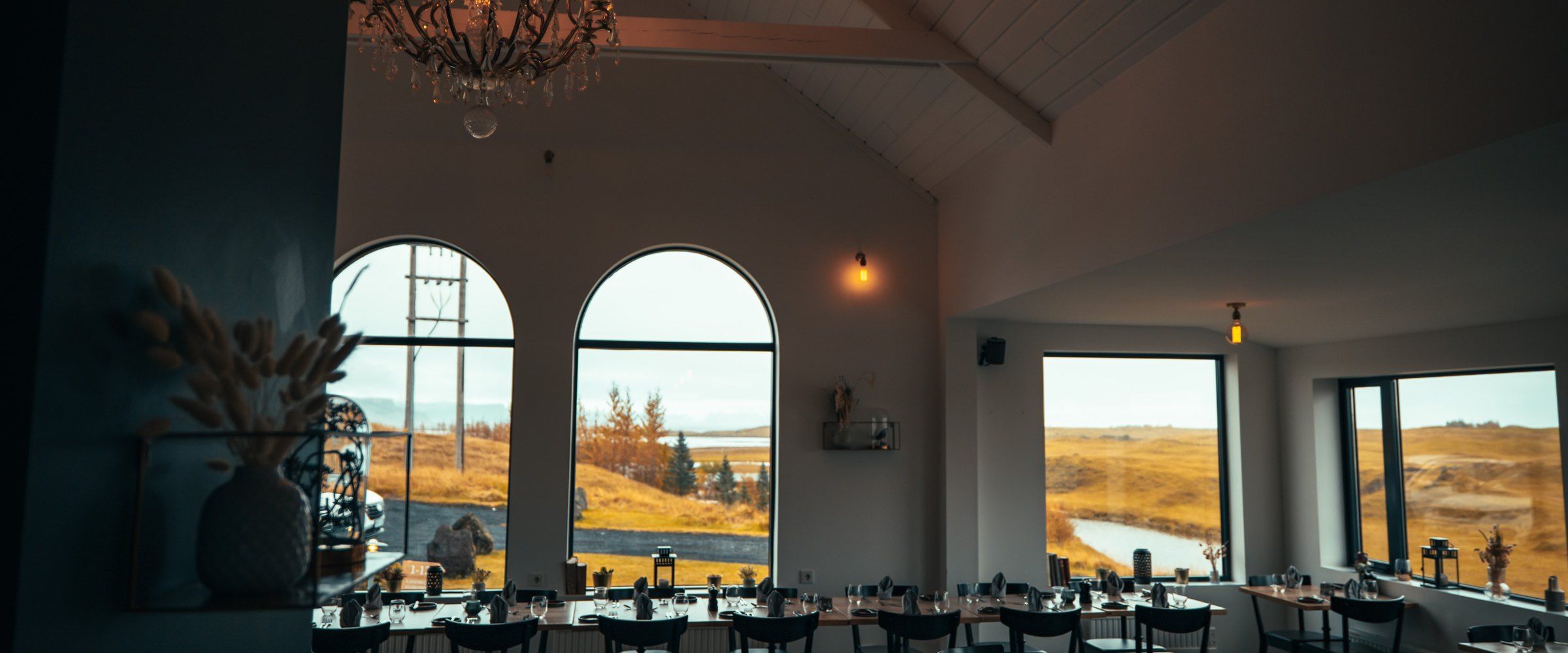 Un comedor con grandes ventanales que ofrecen vistas al paisaje, una araña y una mesa lista para comer.