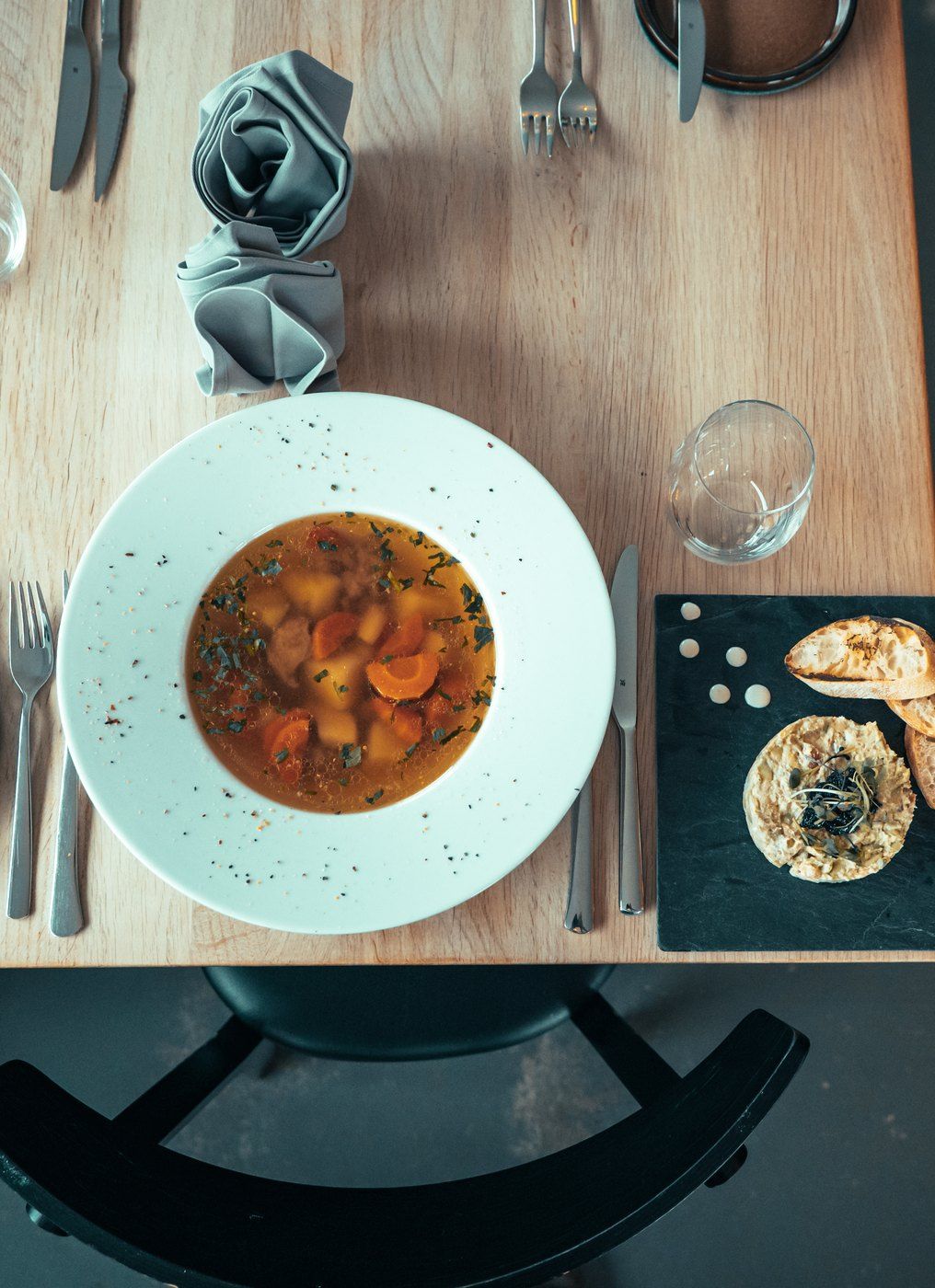 A dining table with a plate of soup, cutlery, a napkin, and a slate board with bread.