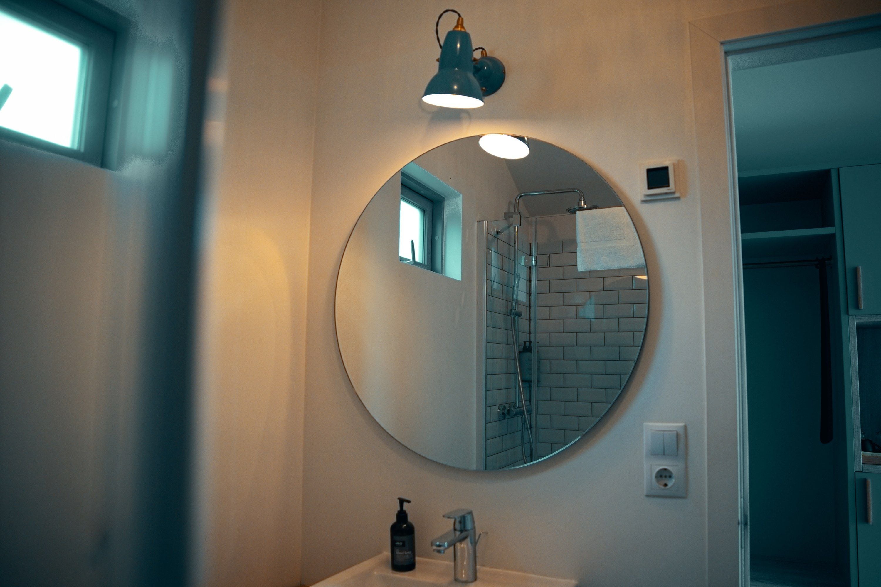 A bathroom with a round mirror, a sink, a handheld showerhead, and a small window.