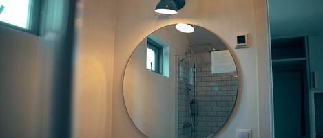 A bathroom with a round mirror, a sink, a handheld showerhead, and a small window.