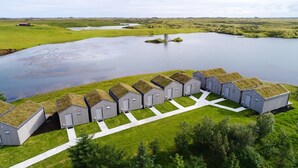Una fila di edifici moderni grigi con tetti verdi, disposti in linea lungo un vialetto erboso accanto a un lago tranquillo.