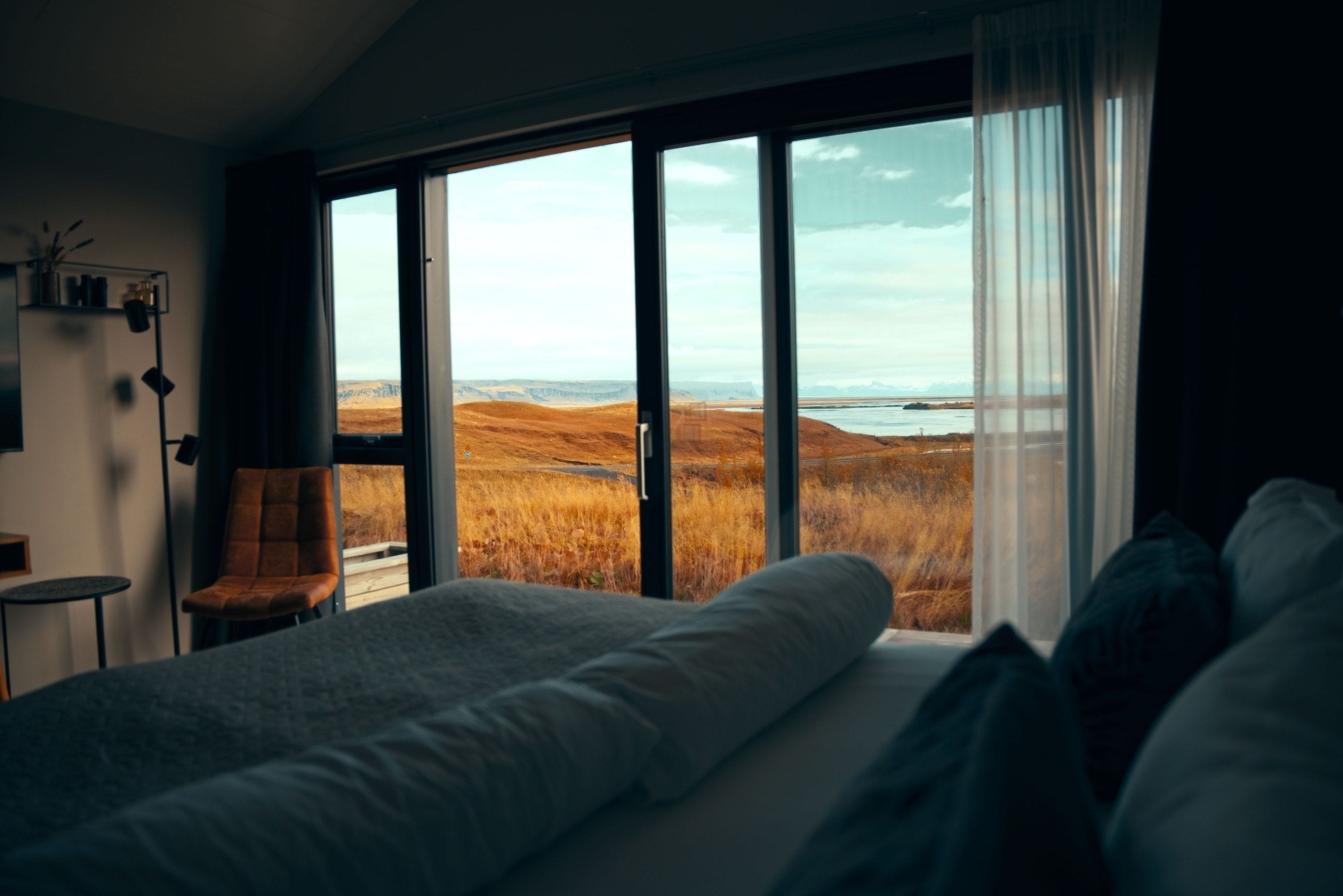 A bedroom with a large bed, a chair, and a view of a landscape through floor-to-ceiling windows.