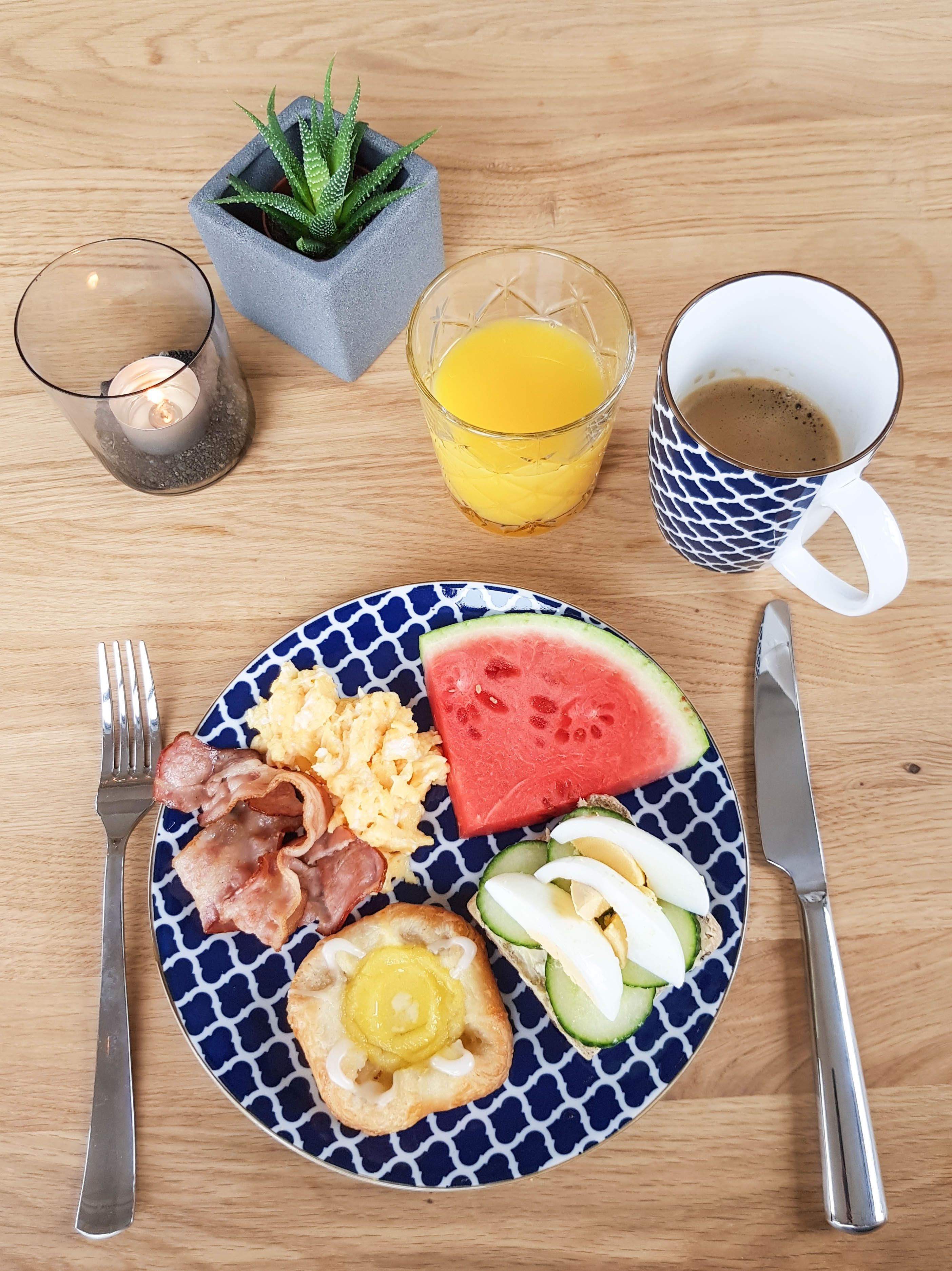 A breakfast plate with scrambled eggs, bacon, a slice of watermelon, and a small pastry, accompanied by a glass of orange juice and a cup of coffee.